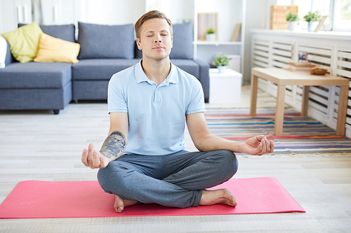 Young man in light blue polo shirt and grey sweatpants sitting on pink mat and practicing yoga exercise