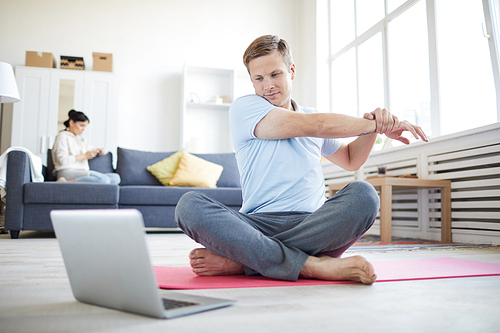 Young man stretching right arm while sitting on the floor with crossed legs and watching online workout in front of laptop