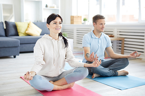 Young active couple sitting in pose of lotus on the floor of living-room during home yoga workout