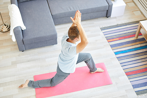 Active young man in shirt and sweatpants exercising on pink mat on the floor of living-room