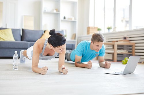 Contemporary young active couple in sportswear doing planks on the floor in front of laptop while watching online workout