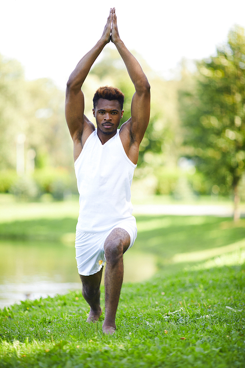 Serious concentrated young African-American man with mustache stretching leg and raising hands in Namaste while strengthening body with yoga exercise in summer park