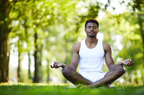 Calm relaxed black man with mustache sitting with crossed legs and holding hands in mudra on knees while meditating alone in summer park