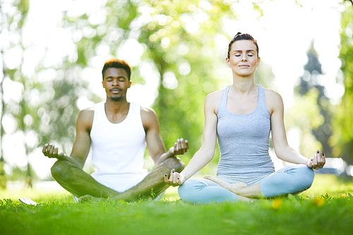 Calm young multi-ethnic man and woman keeping eyes closed and sitting in lotus positions while practicing yoga outdoors