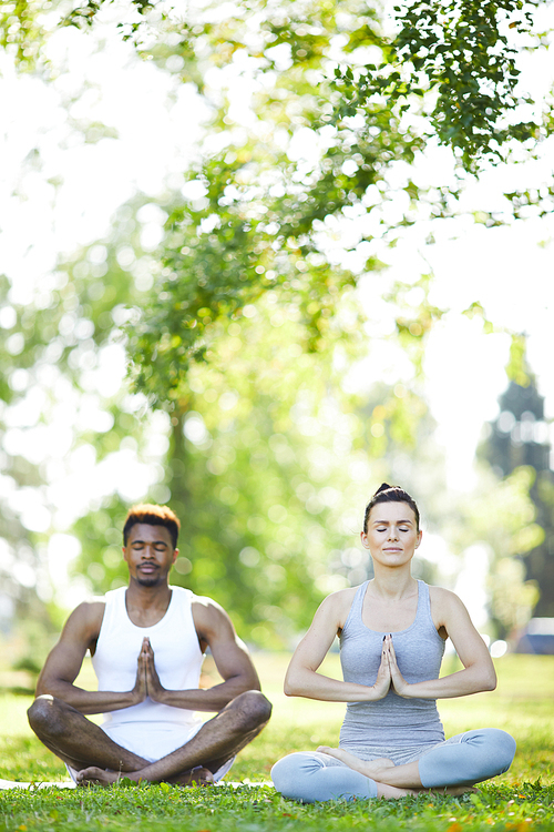 Calm peaceful young multi-ethnic couple sitting with crossed legs on grass and holding hands together in Namaste while meditating in summer park