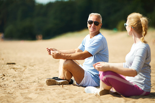 Senior man and his wife in activewear meditating on sand after morning workout