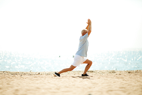 Active retired man in sportswear stretching legs and keeping his raised hands together while exercising on the beach