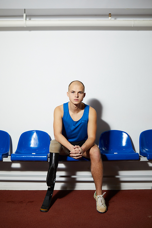 Disable young sportsman with handicap sitting on blue plastic chair along wall before competition