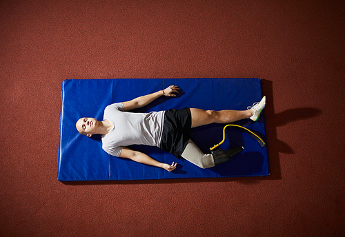 Young relaxed paralympic sportsman with prosthesis on right leg lying on mat before game