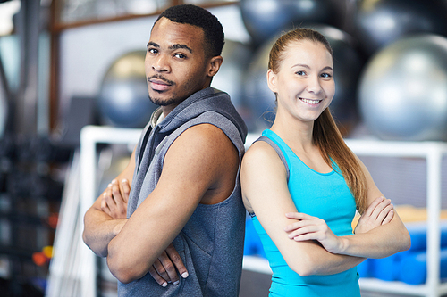 Young sportsman and sportswoman looking at you while standing back to back in gym or fitness center