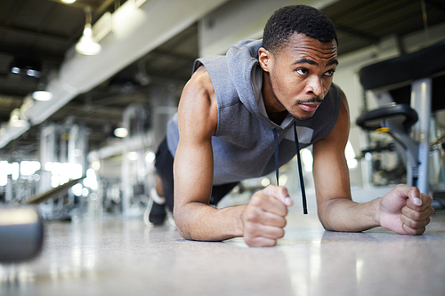 Young African-american man in activewear doing plank on the floor of training gym or fitness center