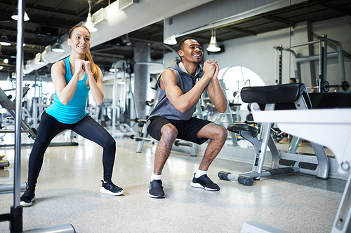 Two young intercultural athletes in activewear doing squats while training in gym or leisure center