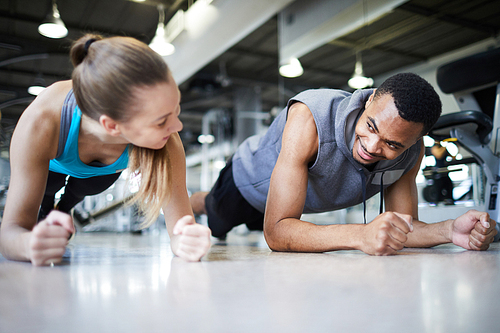 Young intercultural active couple doing plank while looking at one another during workout in gym