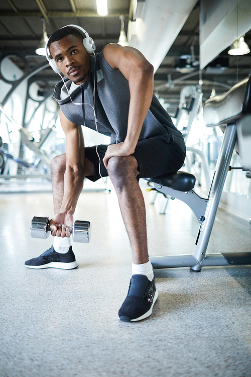 Active African-american guy with dumbbells exercising in gym and listening to his favorite music in headphones