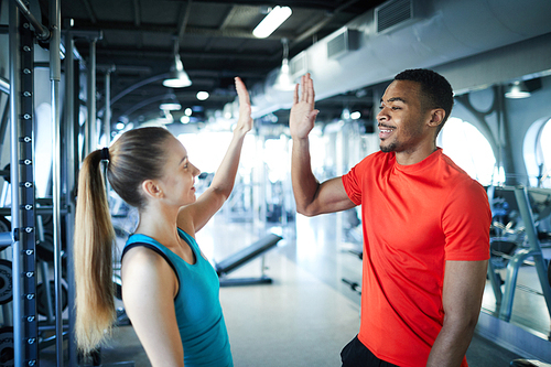 Young intercultural successful trainers giving high five to one another in modern fitness center