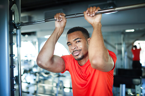 Active African-american athlete in red t-shirt hanging on steel sports bar during workout in modern fitness center