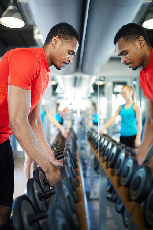 Young athlete taking two heavy barbells from row in front of mirror before exercising in fitness center