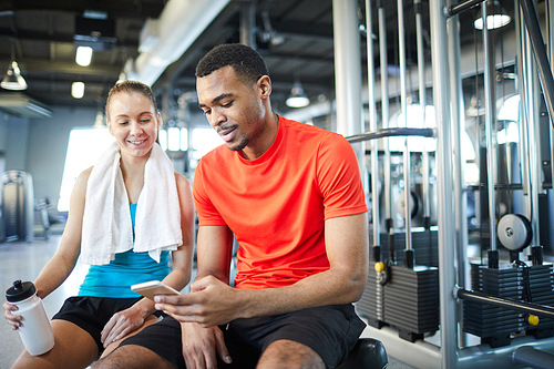 Two intercultural athletes in activewear scrolling through previews in smartphone while having break after workout in gym