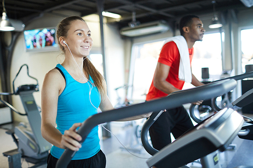 Young active woman running on treadmill and listening to her favorite music in earphones in fitness center