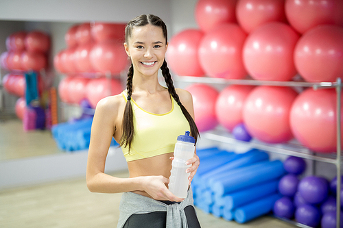 Fit smiling girl with two pigtails holding plastic bottle with water while having break between workouts