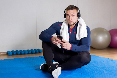 Young athlete in sportswear sitting on mat after training and listening to music in smartphone