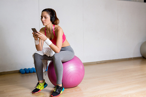 Young restful female in sportswear sitting on fitball at break and listening to tracks from playlist in smartphone
