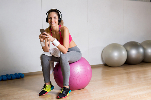 Happy sporty girl with smartphone listening to music while sitting on fitness ball