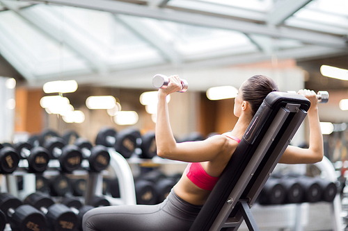 Young active woman sitting on sports equipment and exercising with dumbbells in gym