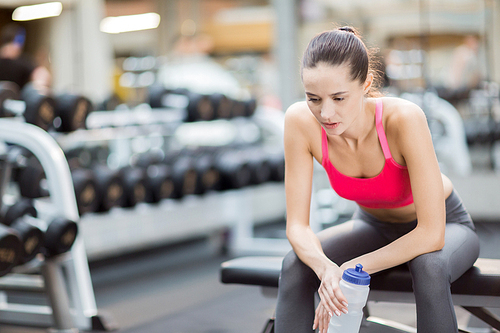Tired young woman with bottle of water sitting on black leather bench in gym after hard training