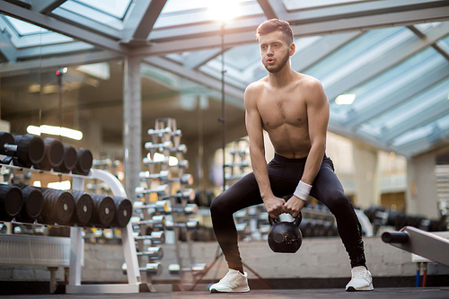 Strong young man doing difficult exercise with heavy kettlebell while lifting it over floor during workout