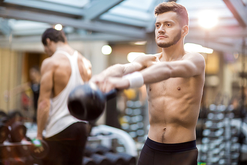 Shirtless guy with stretched arms lifting heavy kettlebell while exercising in modern sports club