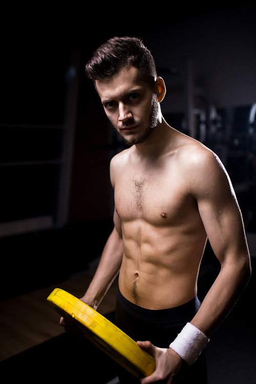 Shirtless sporty guy with heavy yellow metallic disk  while training in cross training center