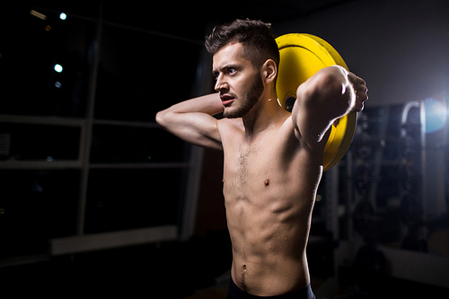 Sporty muscular man holding heavy barbell disc behind head while exercising in gym