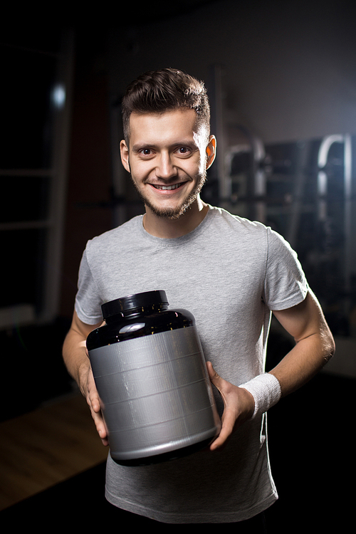 Happy young sportsman holding big plastic jar with protein nutrition