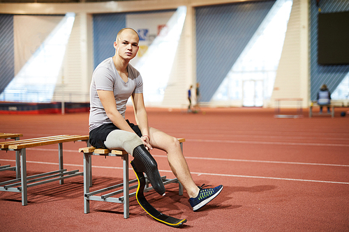 Young paralympic sportsman with prosthesis on right leg sitting on bench at stadium