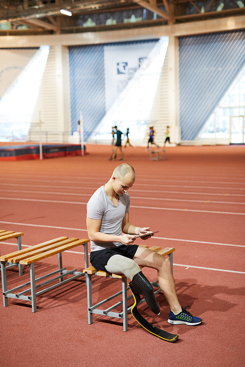 Sportsman with artificial right leg searching in the net while resting on bench by racetracks at stadium