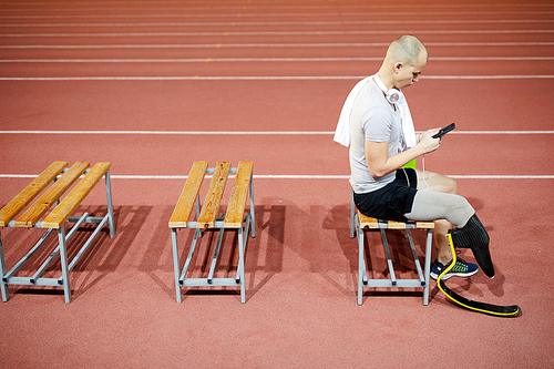 Young sportsman with prosthesis on right leg sitting on bench at break and looking for something to listen in playlist