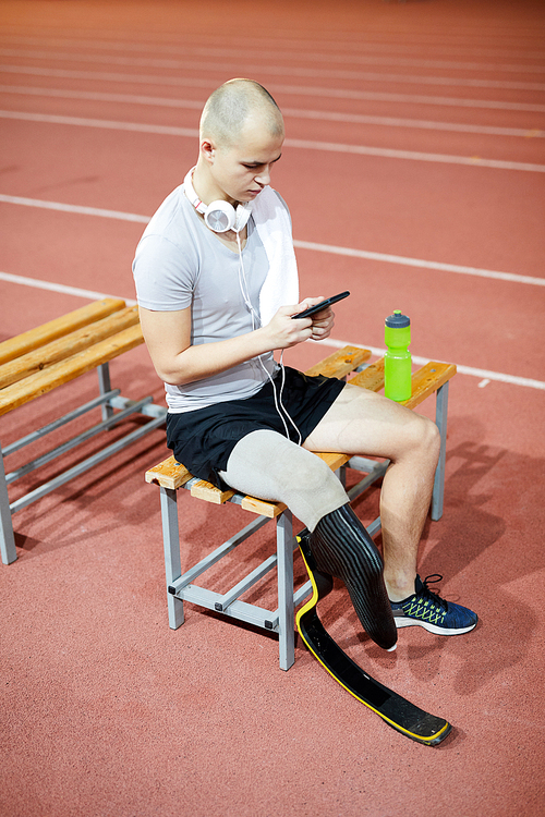 Young paralympic games competitor with smartphone and headphones choosing something to listen from playlist
