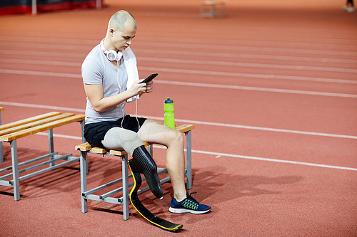 Young sportsman with prosthesis instead of amputeted right leg scrolling in smartphone at break after training on stadium