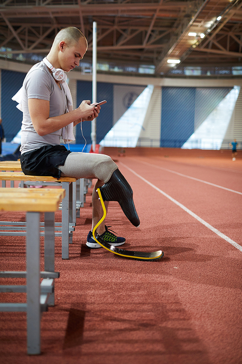 Side view of young paralympic sportsman using smartphone while resting on bench after training at stadium