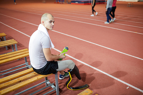 Young active man with handicapped right leg having break and refreshment on bench after training
