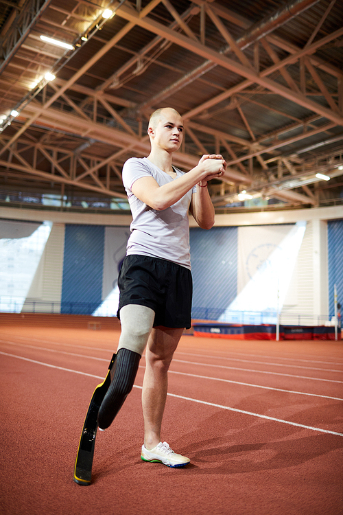 Young paralympic sportsman with prosthesis on right leg doing exercise for hands while standing on racetrack