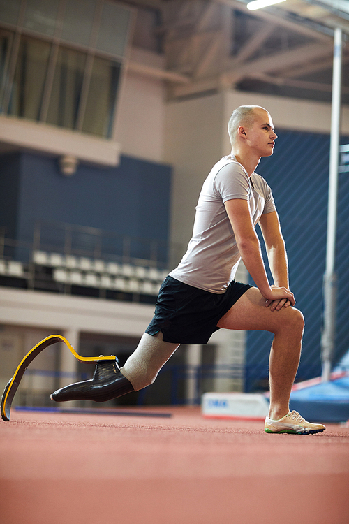 Young active man with prosthesis in right leg doing stretching exercise during workout on stadium
