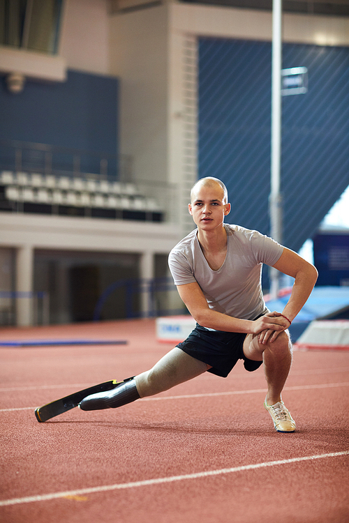 Young man in activewear doing stretching exercise for legs on racetrack on stadium while preparing for paralympic games