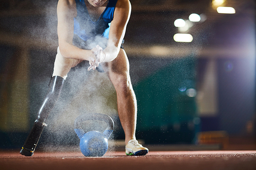 Young paralympic athlete applying talc on his hands while leaning over kettlebell at stadium