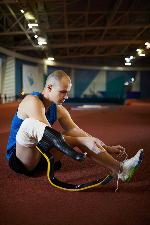 Sportsman with handicapped leg sitting on the floor and tying shoelace of his sneaker before training