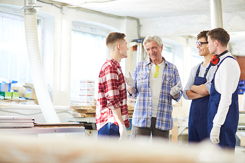 Smiling skilled handsome mature carpenter in checkered shirt standing in circle of young interns and explaining rules to them in modern workshop
