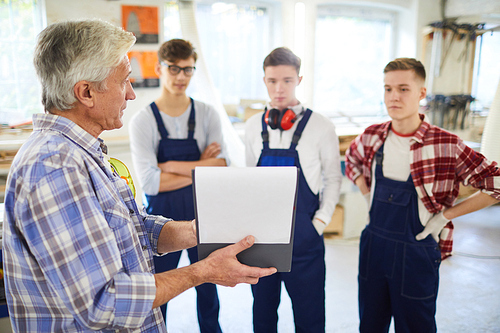 Serious confident mature woodworking master in checkered shirt holding clipboard with papers and showing sketch while explaining project to young students in workshop