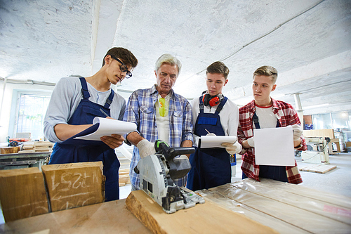 Serious concentrated woodworking specialist with gray hair standing at desk with circular saw and showing young interns with clipboards how to use it