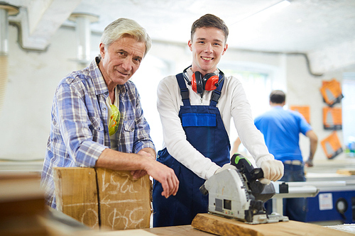 Happy gray-haired mature carpenter and his intern with ear protectors on neck  while standing at workbench with power tool in modern workshop